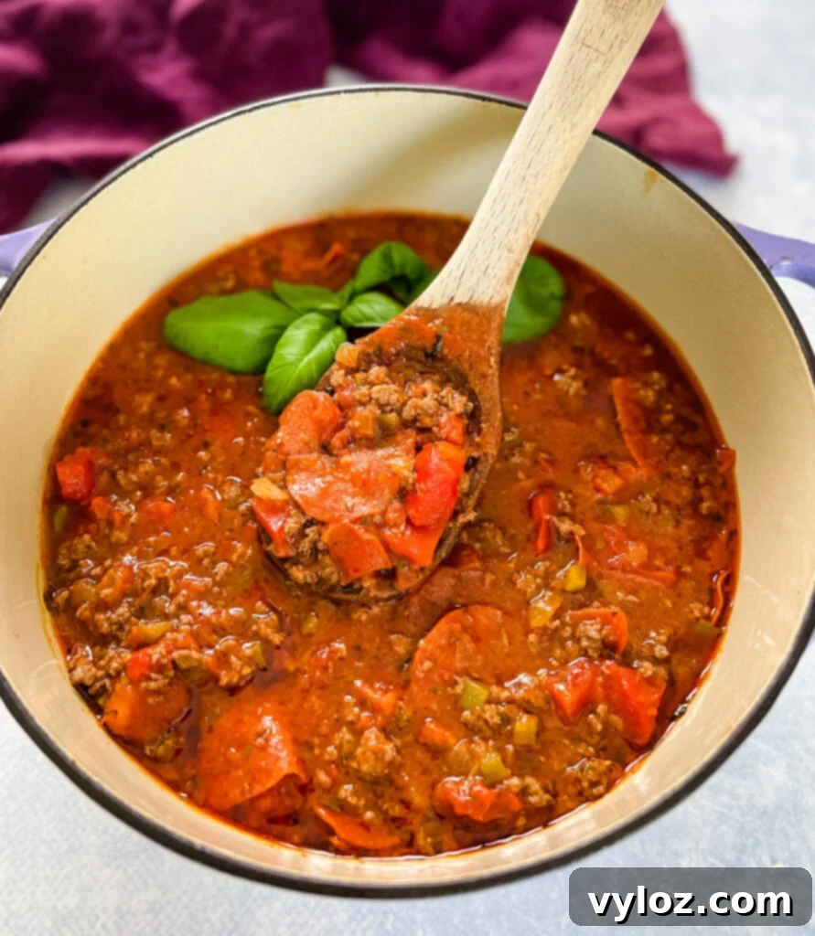 A close-up of hearty pizza soup with beef, pepperoni, and fresh basil simmering in a dark pot, ready to be served