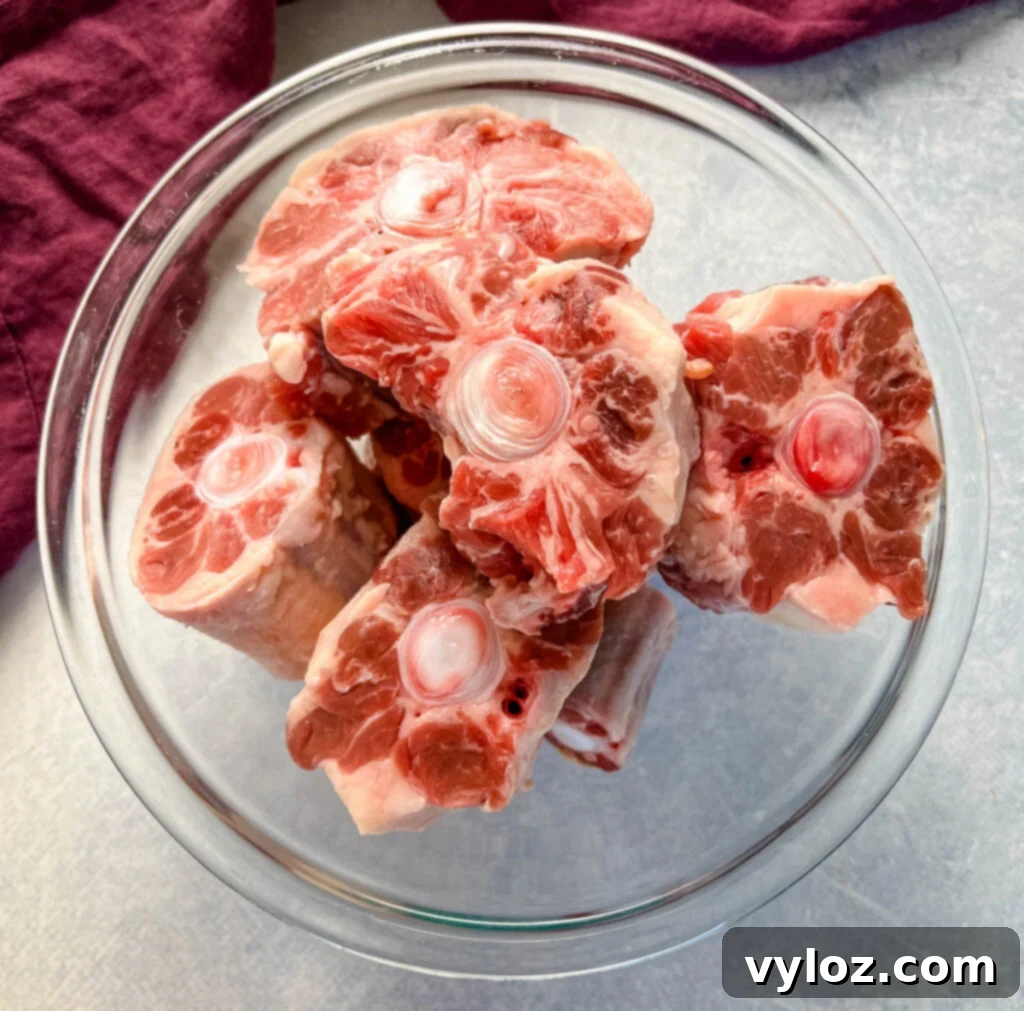 Uncooked, trimmed oxtail pieces in a clear glass bowl, showcasing the marbled meat and bone structure before seasoning