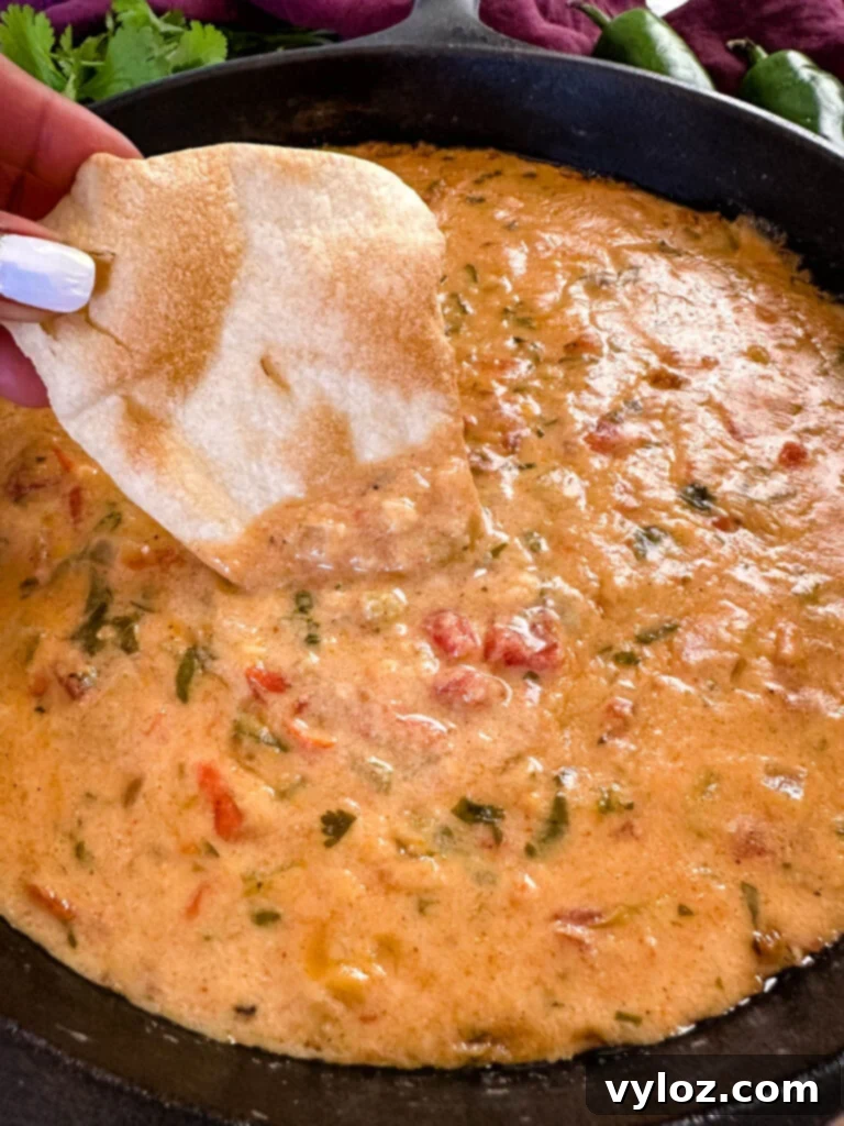 A person holding a chip dipped in smoked queso cheese dip, ready to eat, with the skillet in the background