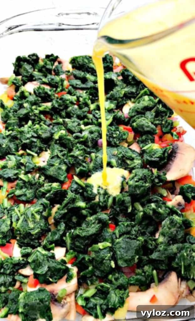 Raw egg being poured into a dish filled with various vegetable ingredients for a Low-Carb Bacon, Egg, and Spinach Breakfast Casserole in a glass pan