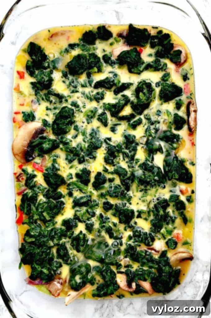 Raw egg mixture combined with vegetable ingredients for Low-Carb Bacon, Egg, and Spinach Breakfast Casserole in a glass pan, ready for the next step
