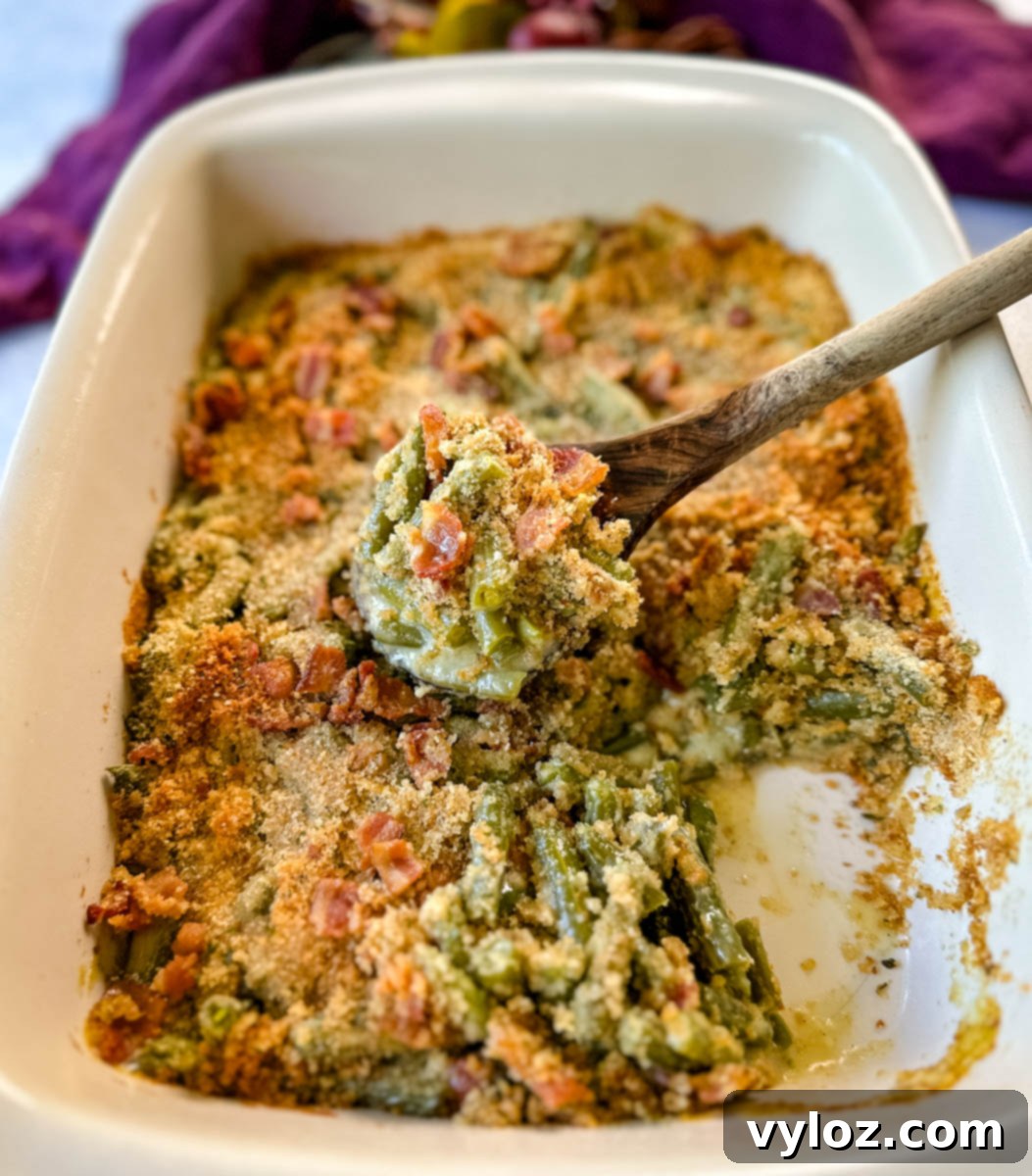 A wooden spoonful of green bean casserole with bacon and breadcrumbs being served from a baking dish