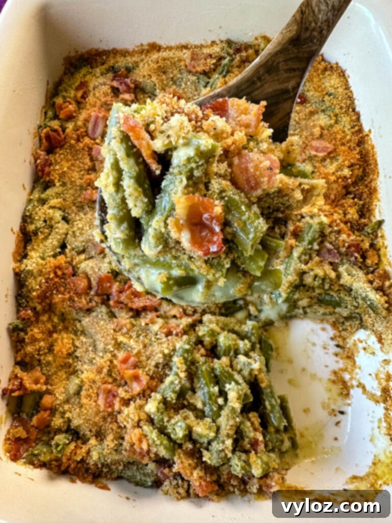 A wooden spoonful of green bean casserole with bacon and breadcrumbs in a baking dish