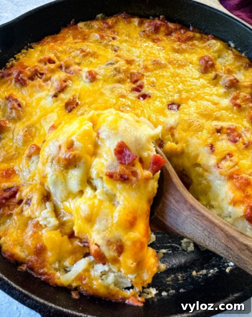 A wooden spoonful of cheesy bacon hash brown casserole being lifted from a warm cast iron skillet.