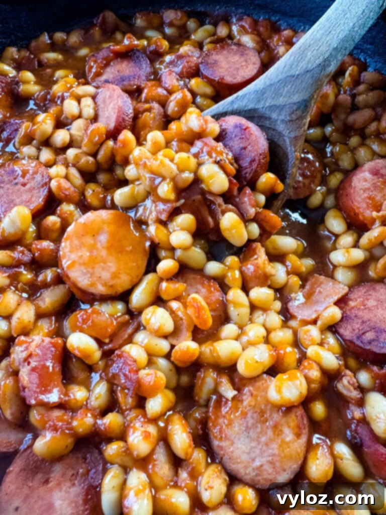 homemade pork and beans with sausage, bacon, and a wooden spoon in a cast iron skillet
