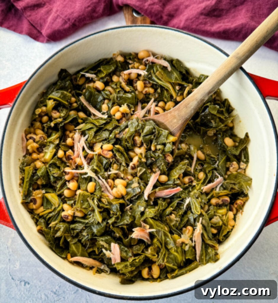 A top-down view of finished collard greens and black-eyed peas, enriched with smoked turkey, presented in a red Dutch oven with a wooden spoon for serving.