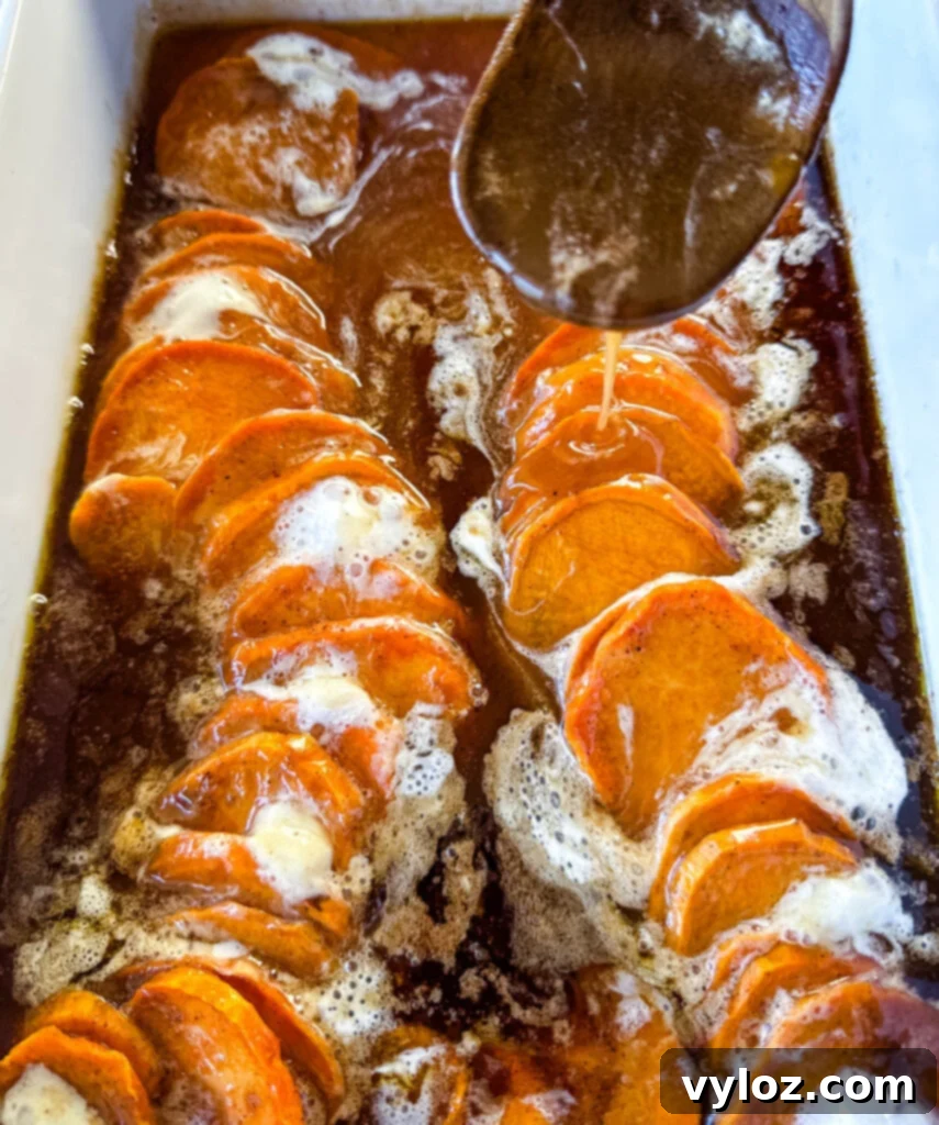 Classic candied sweet potatoes with toasted marshmallows in a baking dish, with a wooden spoon drizzling extra glaze.