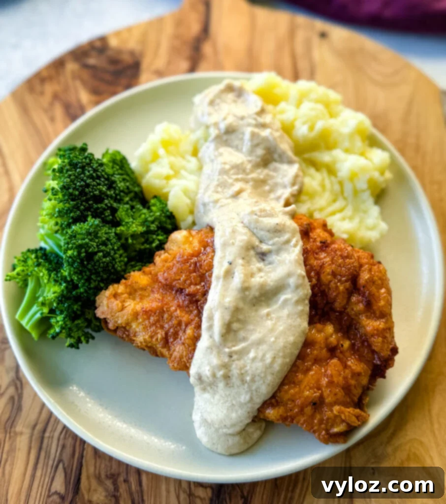 Crispy chicken fried chicken with creamy country gravy, mashed potatoes, and broccoli on a plate, ready to be enjoyed.