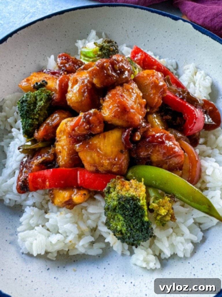 A serving of orange chicken with stir-fried vegetables and white rice in a white bowl, ready for enjoyment.