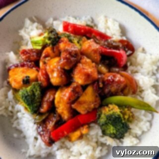 Orange chicken with stir-fried vegetables and white rice in a white bowl.