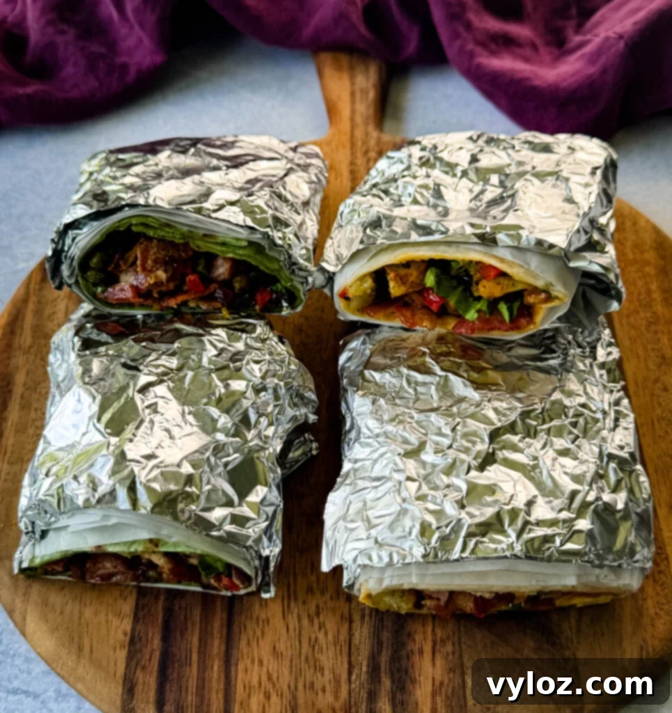 breakfast burritos wrapped with parchment paper and foil with bacon, potatoes, bell peppers and cheese on a flat surface