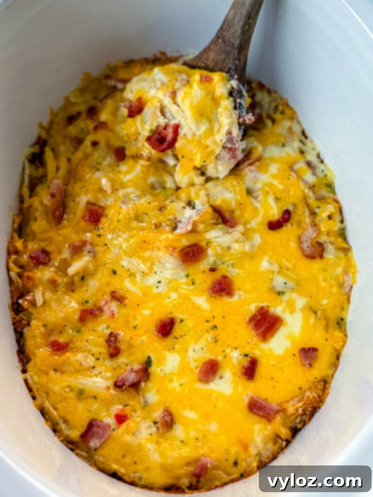 A wooden spoonful of golden-brown hash brown casserole, rich with melted cheese and savory bacon, held above a Crockpot slow cooker, ready to be served.