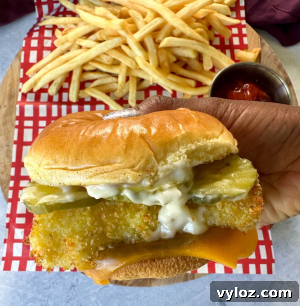 Golden Fried Fish Sandwich 11 person holding fish sandwich with cheese, pickles, and tartar sauce on a brioche bun on a plate with fries