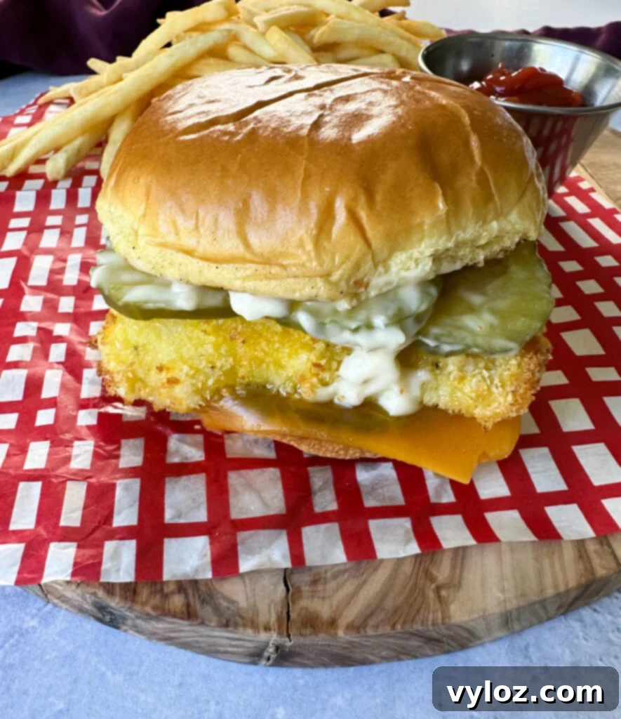 Golden Fried Fish Sandwich 10 fish sandwich with cheese, pickles, and tartar sauce on a brioche bun on a plate with fries