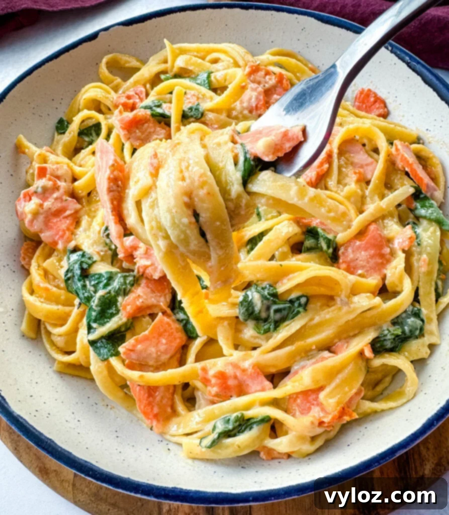 A fork lifting a generous portion of creamy Salmon Fettuccine Alfredo with spinach from a white bowl.