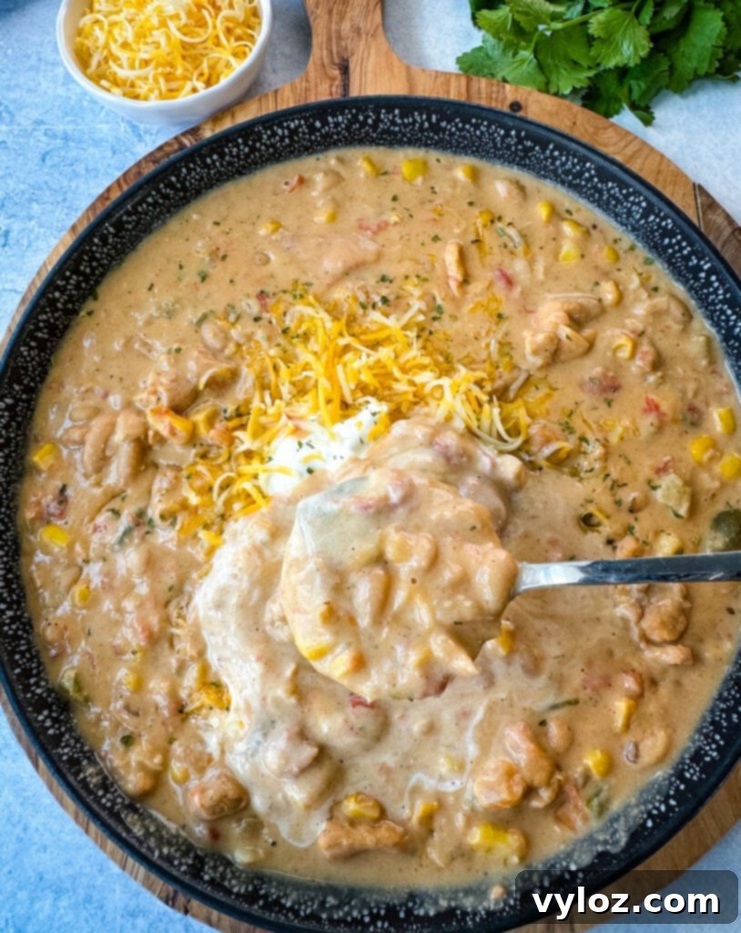 A black bowl of white chicken chili with a spoon, generously topped with sour cream and grated cheese, against a rustic background.