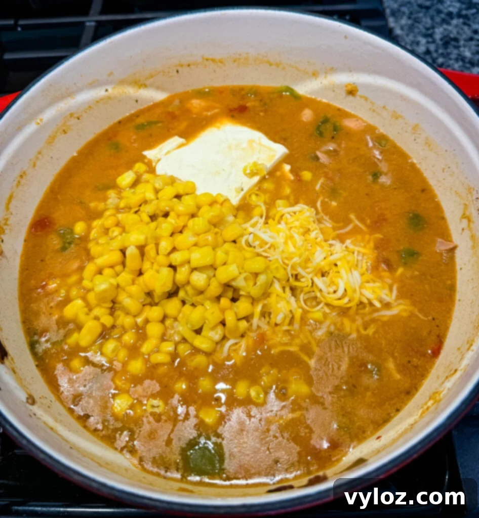 White chicken chili simmering in a Dutch oven, with corn, grated cheese, and cream cheese visible before being fully stirred in, ready to create a creamy texture.