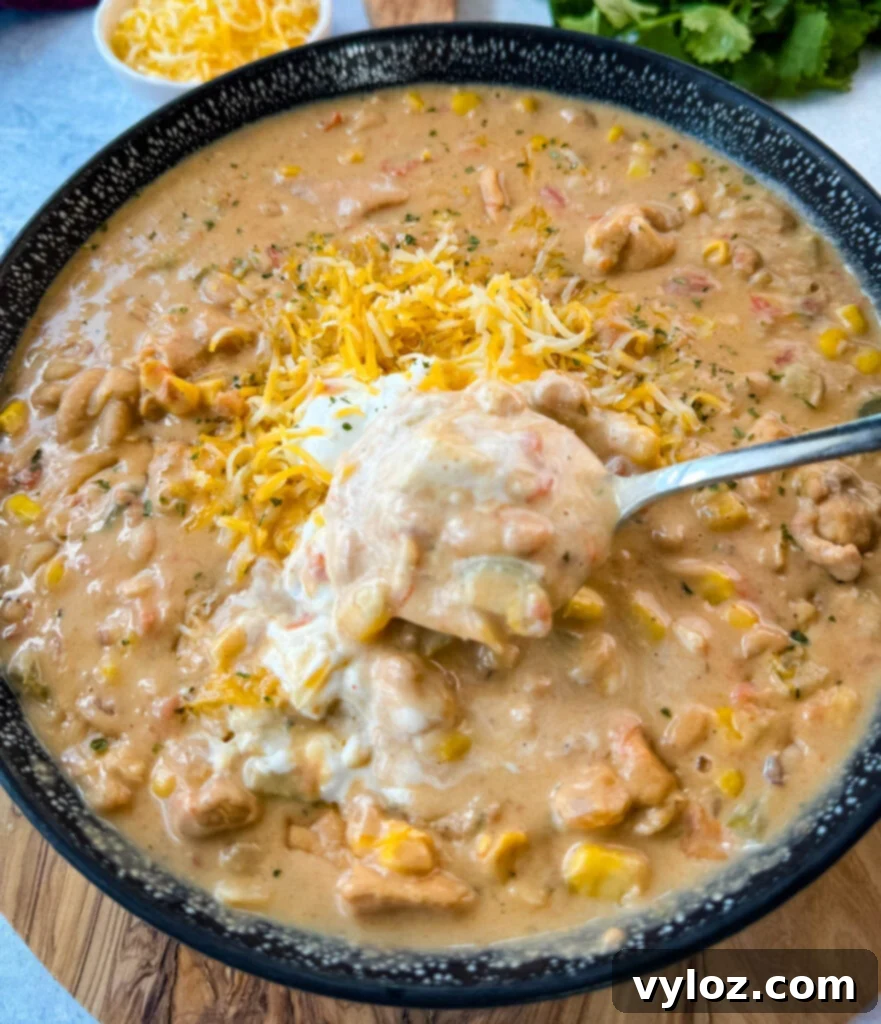 A black bowl of white chicken chili, garnished with sour cream and grated cheese, with a spoon resting inside, invitingly ready to be eaten.