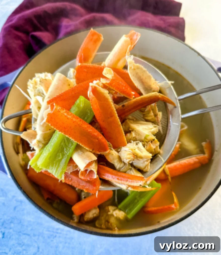Oceanic Flavor Base 7 A fine-mesh strainer holding cooked crab shells, carrots, and celery over a Dutch oven filled with clear, golden homemade seafood stock.