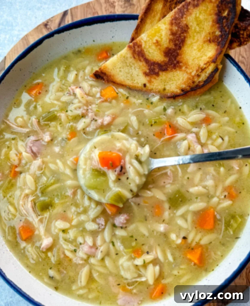 chicken orzo soup with carrots, celery, and a wooden spoon in a white bowl with a piece of bread and a spoon