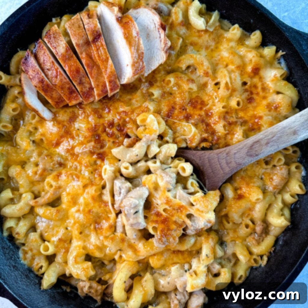 A serving of chicken mac and cheese in a cast iron skillet, garnished with sliced chicken and a wooden spoon, ready to serve.