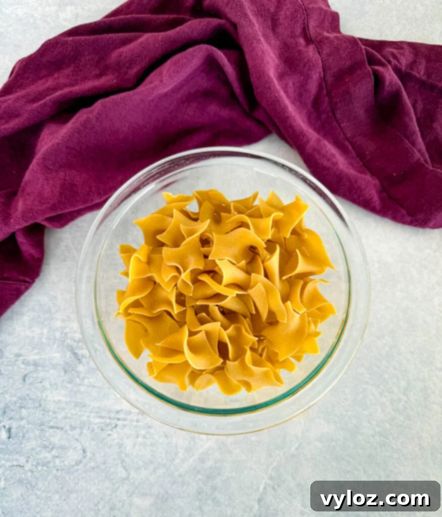 Dry wide egg noodles piled neatly in a clear glass bowl, showcasing their texture and readiness for cooking.