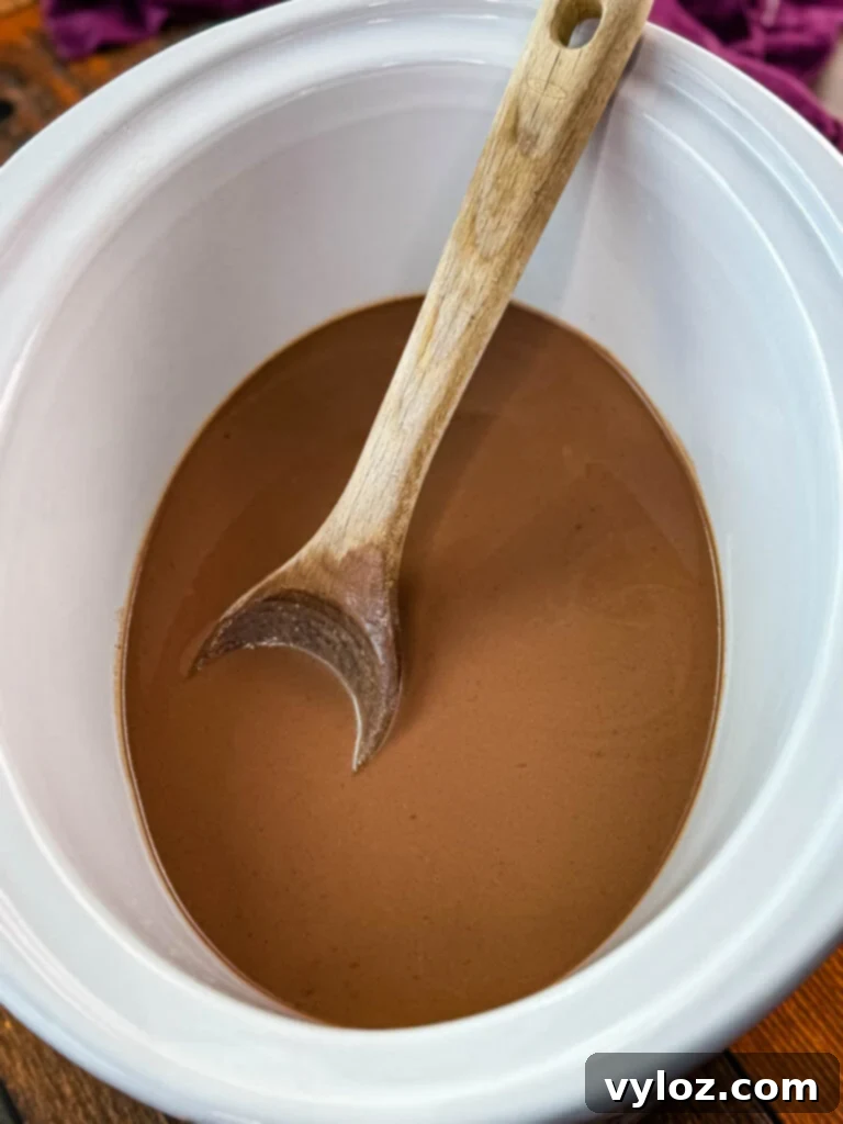 Hot chocolate simmering in a Crockpot slow cooker with a wooden spoon, showcasing its smooth texture.