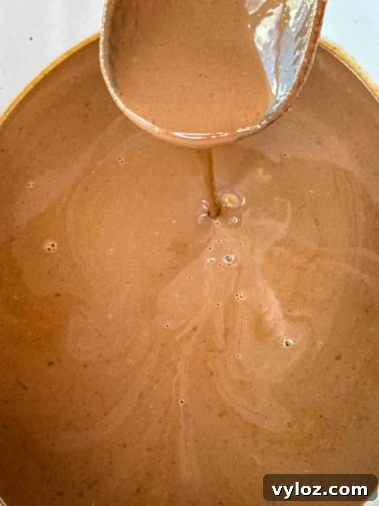 Rich, dark hot chocolate being stirred with a wooden spoon in a Crockpot slow cooker, illustrating its creamy consistency.