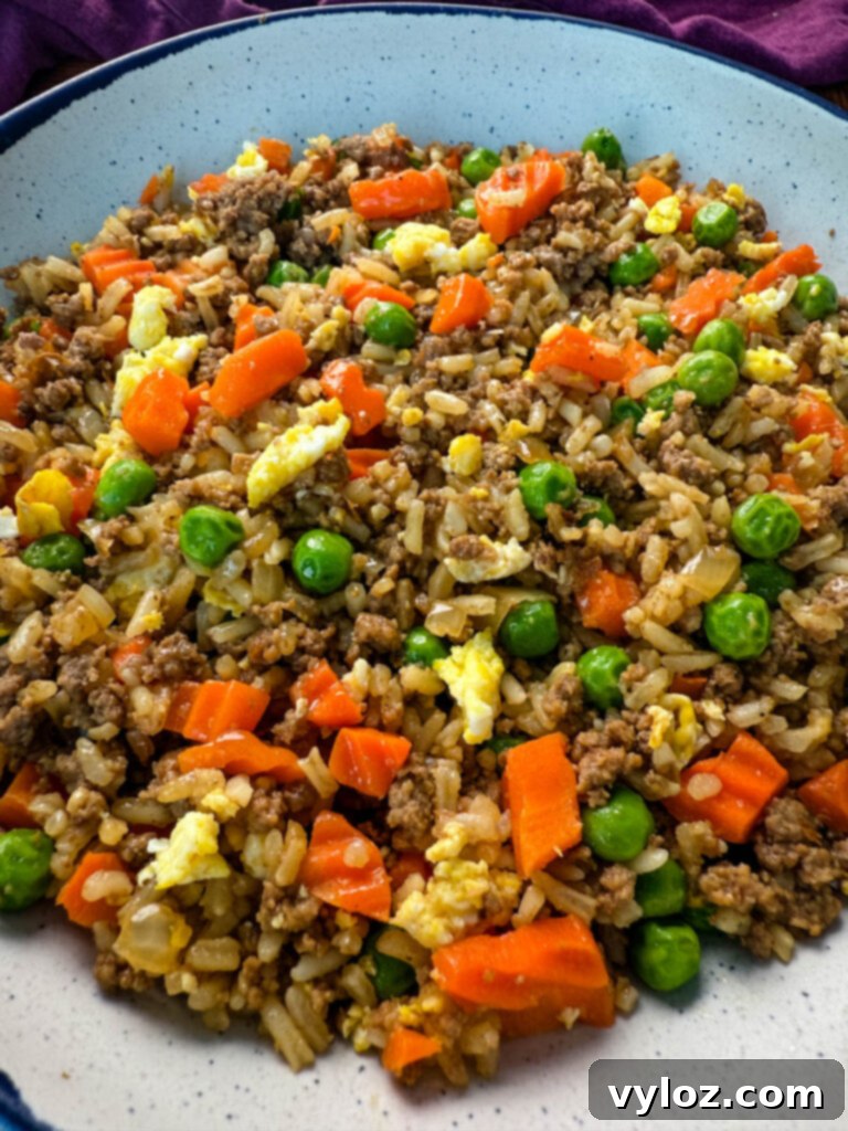 A serving of delicious ground beef fried rice in a white bowl, garnished with scrambled eggs, bright green peas, and diced carrots.
