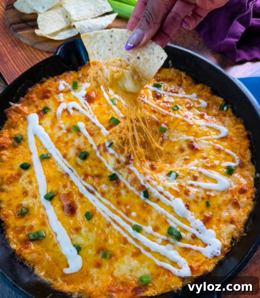 Golden, bubbly Ranch Buffalo Chicken Dip with Rotisserie Chicken in a skillet, topped with drizzled ranch dressing and chopped green onions. A tortilla chip is being dipped, showing the gooey, cheesy pull.