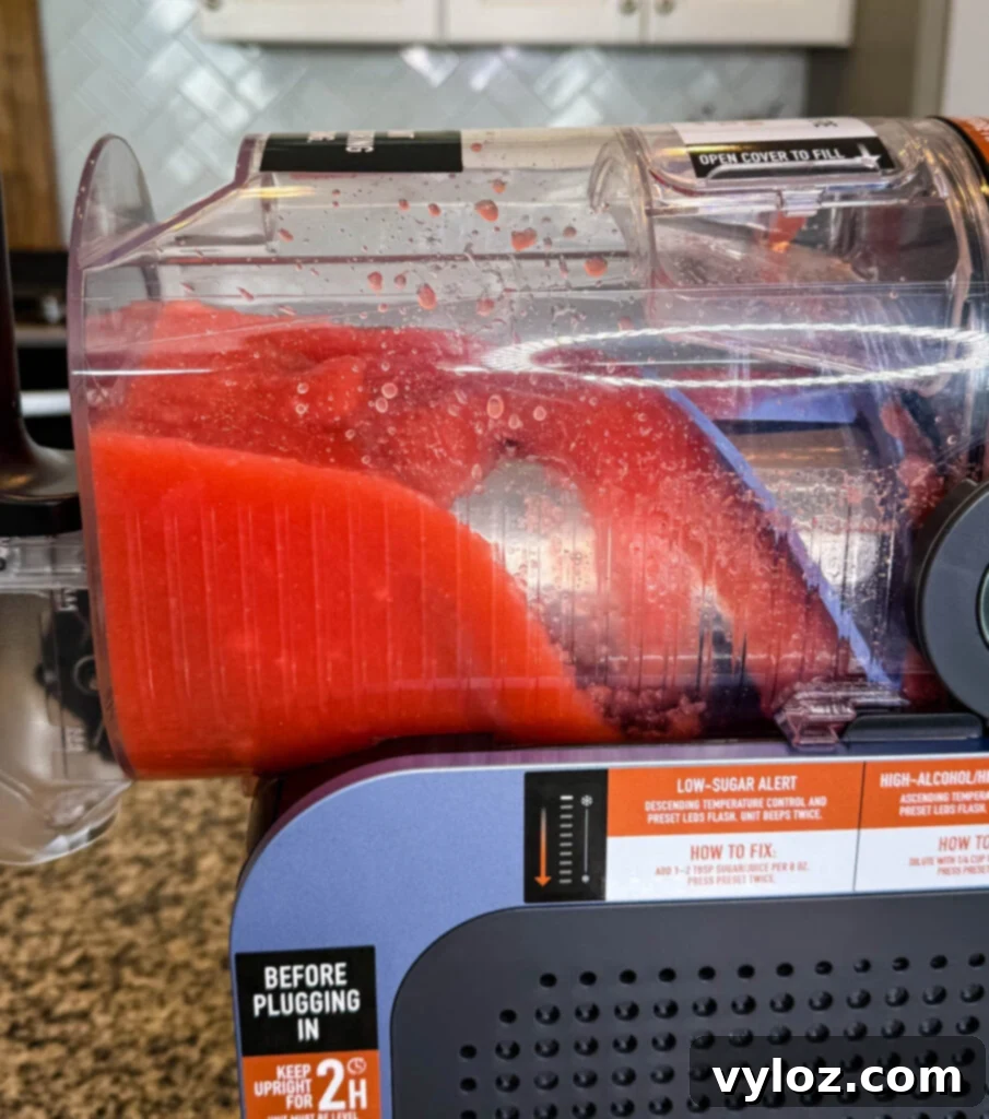 Side View of the Ninja SLUSHi Machine in Action – A side angle of the machine with red slush forming inside the chamber. The condensation and mixing process are visible, along with labels for low-sugar and high-alcohol warnings.