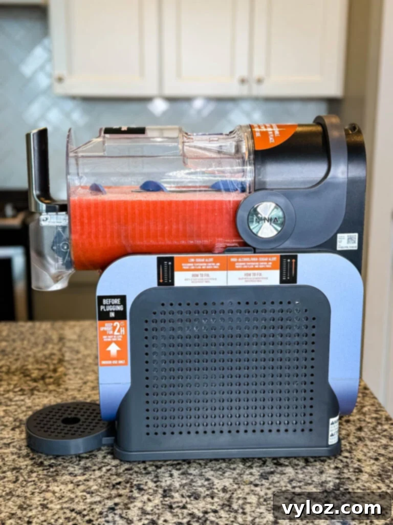 Full Side View of the Ninja SLUSHi Machine on a Kitchen Counter – The machine is placed on a kitchen counter with the chamber filled with a frozen drink. Labels on the side provide usage instructions, including a reminder to keep the unit upright for 2 hours before first use.