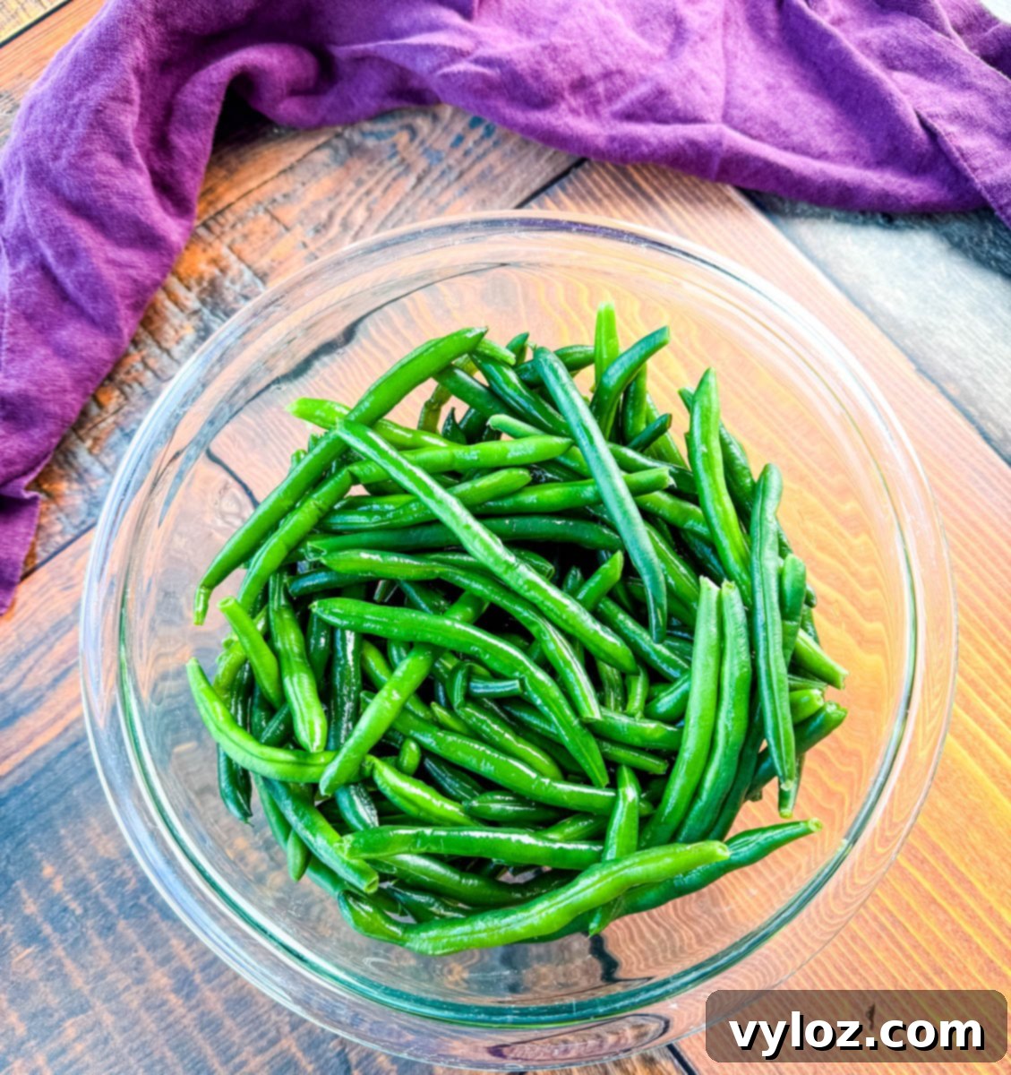 A clear glass bowl filled with bright green cooked green beans on a rustic wooden table with a purple napkin — fresh and ready for Southern green bean casserole.