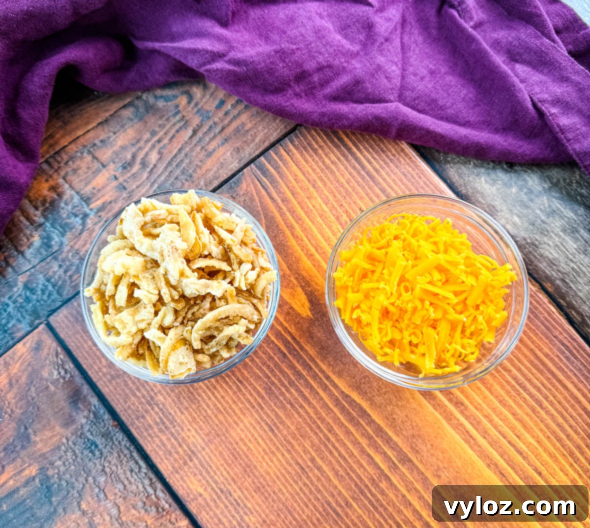 A small glass bowl of crispy fried onions and a bowl of shredded cheddar cheese sit on a wooden surface with a deep-purple napkin in the background — ingredients for Southern-style green bean casserole.