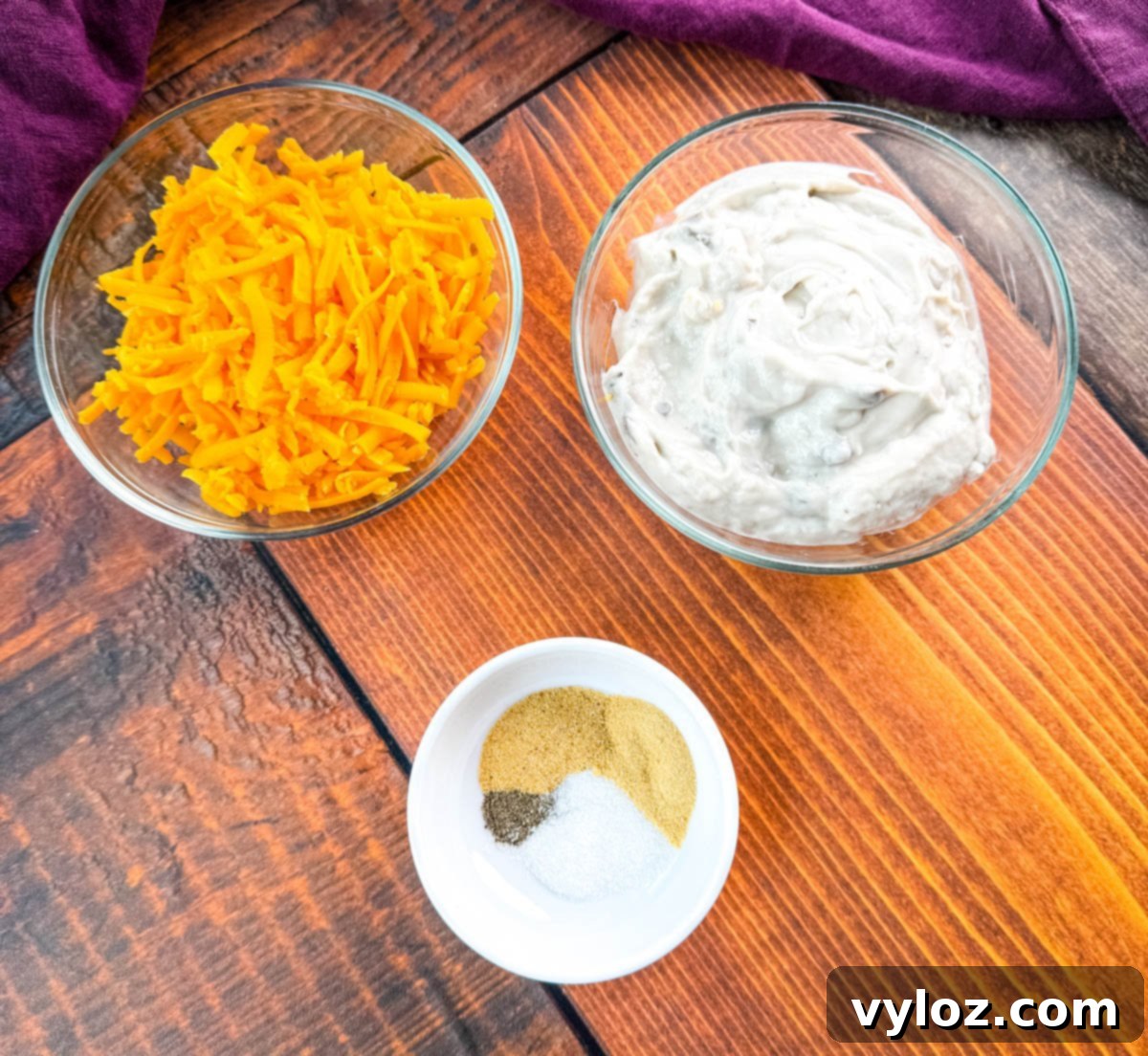 Glass bowls filled with shredded cheddar cheese and creamy mushroom soup, next to a small white dish of seasoning mix including garlic powder, onion powder, salt, and pepper on a wooden background — prep setup for green bean casserole.