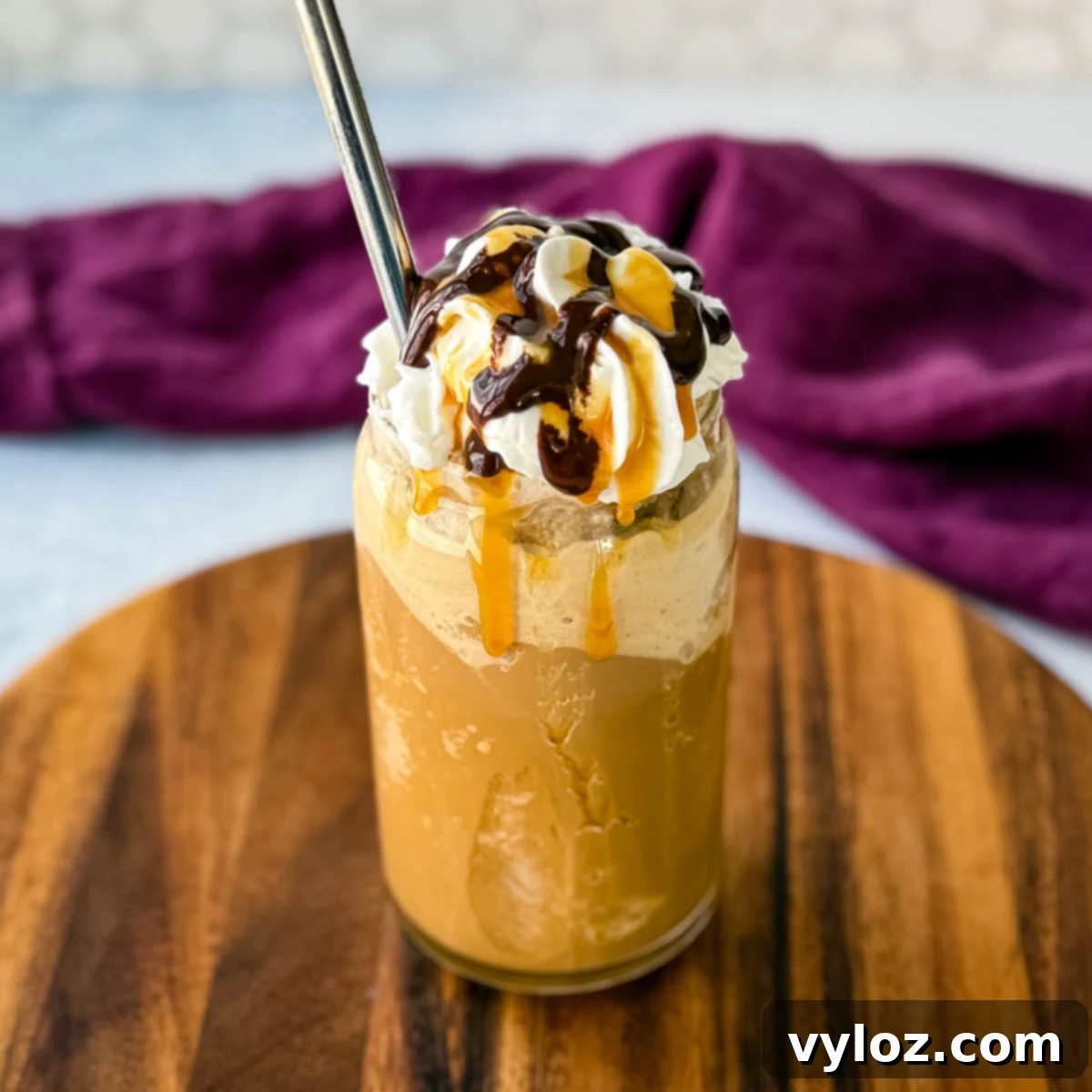 Shadow Swirl Vanilla Caramel Frappé 2 A tall glass filled with a blended coffee frappe, topped with whipped cream, chocolate drizzle, and caramel sauce. A metal straw is inserted, and the glass sits on a wooden board with a purple cloth in the background.
