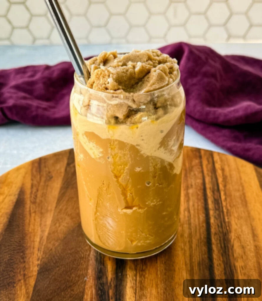 Shadow Swirl Vanilla Caramel Frappé 7 A glass filled with a blended coffee frappe topped with a thick, icy coffee mixture. A metal straw is inserted, and the glass sits on a wooden board with a purple cloth in the background.