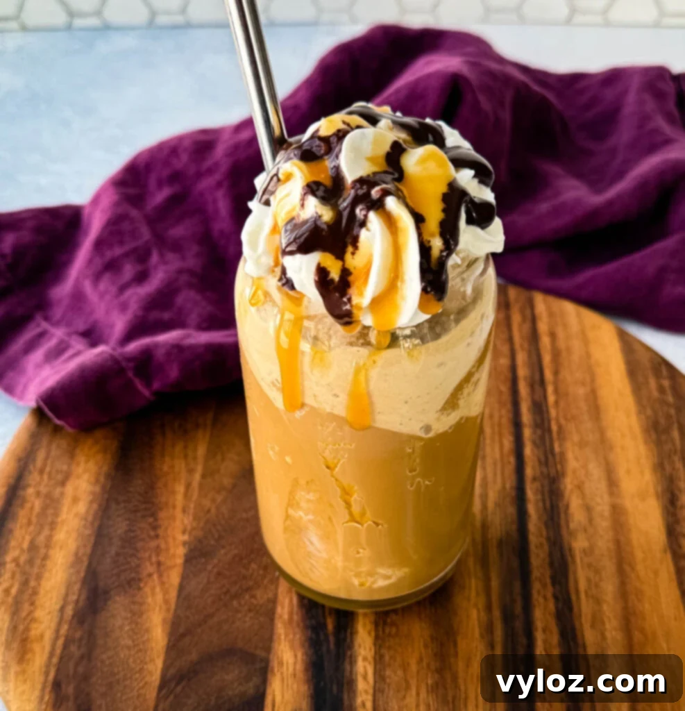 Shadow Swirl Vanilla Caramel Frappé 8 A tall glass filled with a blended coffee frappe, topped with whipped cream, chocolate drizzle, and caramel sauce. A metal straw is inserted, and the glass sits on a wooden board with a purple cloth in the background.