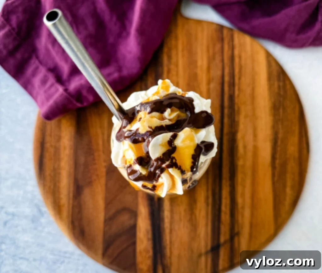 Shadow Swirl Vanilla Caramel Frappé 9 A top-down view of a coffee frappe in a glass, covered with whipped cream, chocolate drizzle, and caramel sauce. A metal straw is inserted, and the drink is placed on a wooden board with a purple cloth partially visible.