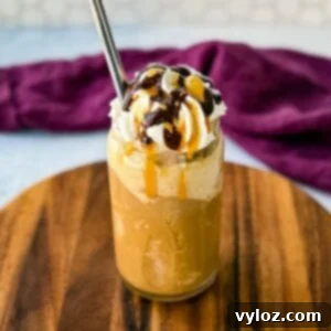 Shadow Swirl Vanilla Caramel Frappé 10 A tall glass filled with a blended coffee frappe, topped with whipped cream, chocolate drizzle, and caramel sauce. A metal straw is inserted, and the glass sits on a wooden board with a purple cloth in the background.