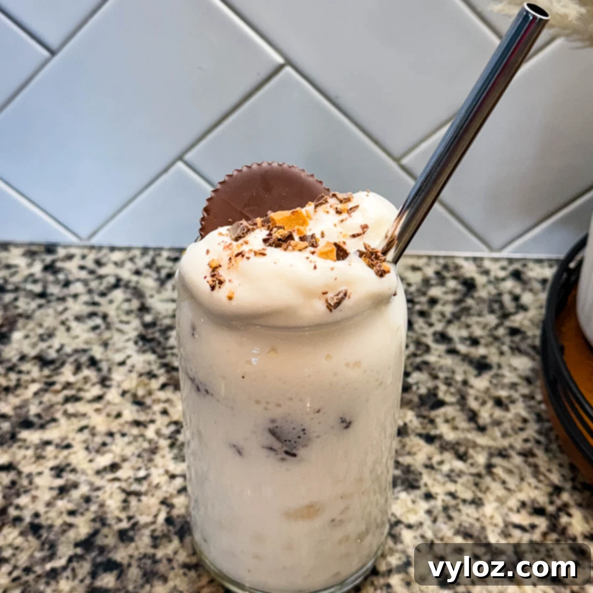 Ninja Slushi Ultimate Vanilla Peanut Butter Cup Protein Milkshake 2 Finished Ninja SLUSHi Peanut Butter Cup Vanilla Protein Shake in a glass, topped with whipped cream, crushed peanut butter cup pieces, and a whole peanut butter cup, with a metal straw for serving.