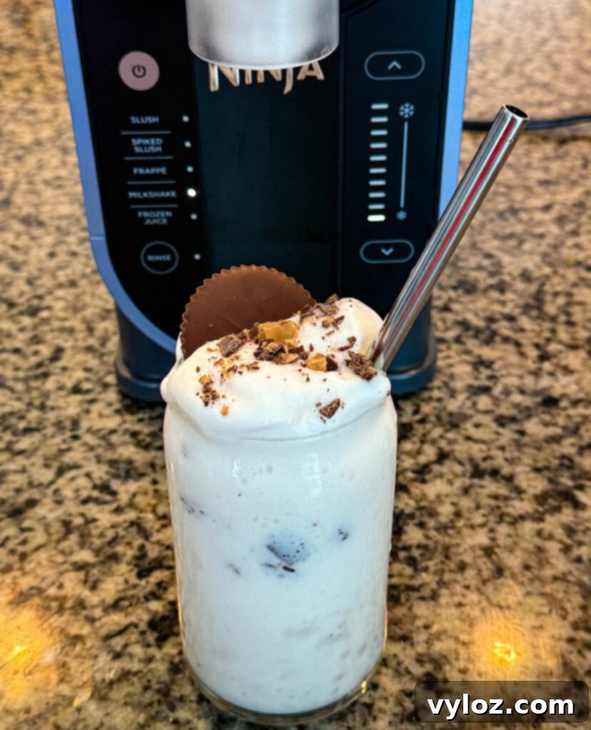 Ninja Slushi Ultimate Vanilla Peanut Butter Cup Protein Milkshake 6 Finished Ninja SLUSHi Peanut Butter Cup Vanilla Protein Shake in a glass, topped with whipped cream, crushed peanut butter cup pieces, and a whole peanut butter cup, with a metal straw for serving.