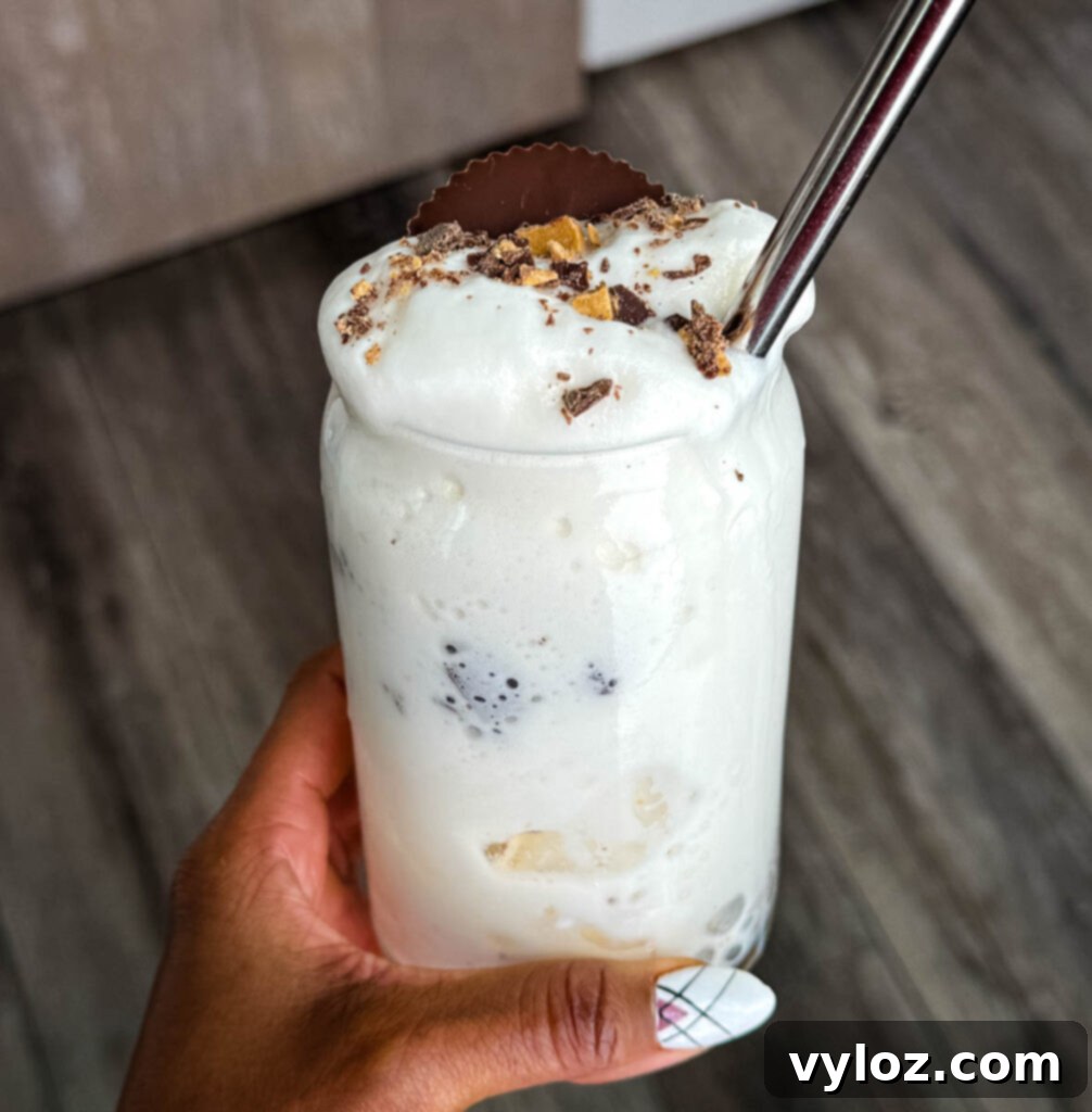 Ninja Slushi Ultimate Vanilla Peanut Butter Cup Protein Milkshake 7 person holding Finished Ninja SLUSHi Peanut Butter Cup Vanilla Protein Shake in a glass, topped with whipped cream, crushed peanut butter cup pieces, and a whole peanut butter cup, with a metal straw for serving.