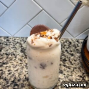 Ninja Slushi Ultimate Vanilla Peanut Butter Cup Protein Milkshake 9 Finished Ninja SLUSHi Peanut Butter Cup Vanilla Protein Shake in a glass, topped with whipped cream, crushed peanut butter cup pieces, and a whole peanut butter cup, with a metal straw for serving.
