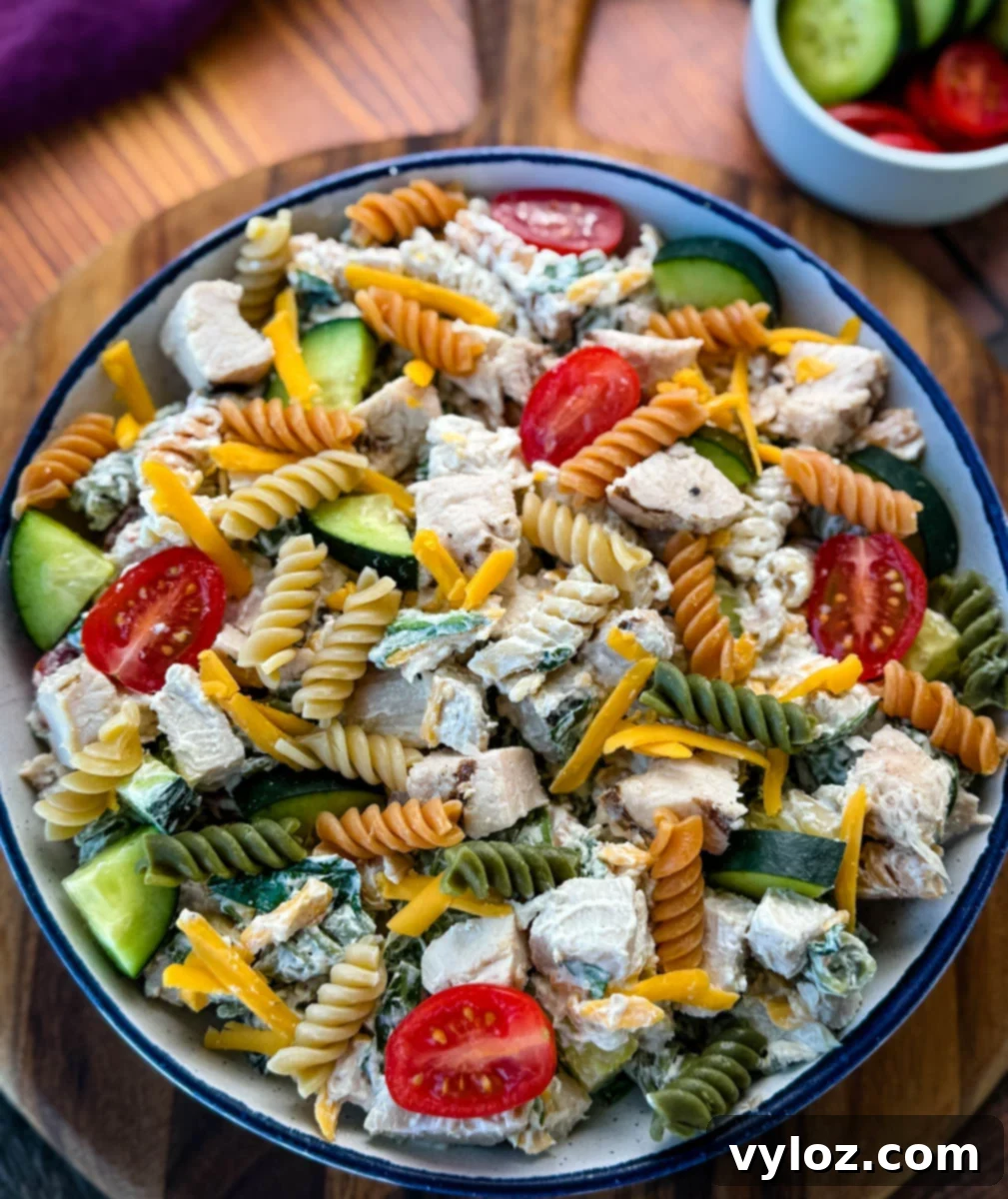Overhead shot of High-Protein Chicken Pasta Salad in a ceramic bowl, garnished with cherry tomatoes, cucumber slices, and cheddar cheese, resting on a wooden backdrop.