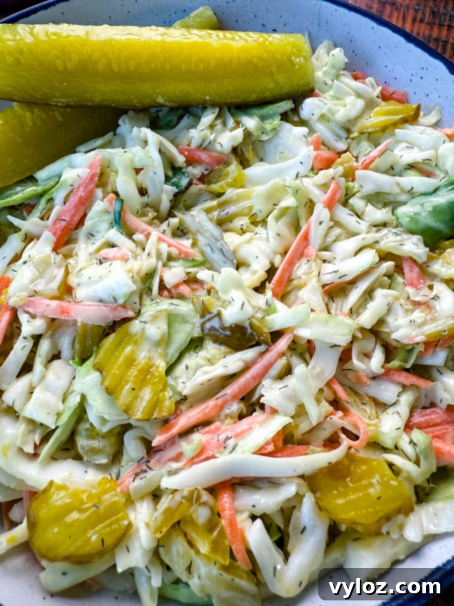 Pickle Lovers' Dream Coleslaw 6 Bowl of Dill Pickle Coleslaw with Pickle Spears on the Side – A close-up of a creamy dill pickle coleslaw served in a rustic bowl, featuring shredded cabbage, carrots, and sliced pickles, garnished with two large dill pickle spears. A small dish of extra pickle slices sits in the background on a wooden surface.