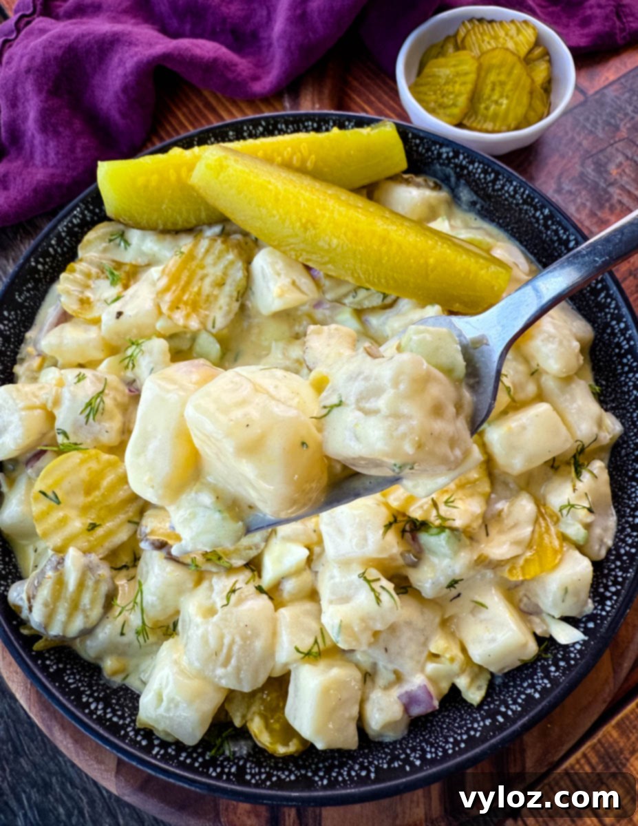 Dill-icious Creamy Potato Salad 11 A spoon lifting a bite of creamy dill pickle potato salad, showcasing the texture of the cubed potatoes, crunchy pickles, and fresh dill. The bowl of salad sits on a wooden surface with a small dish of pickles in the background, creating a visually appetizing presentation.
