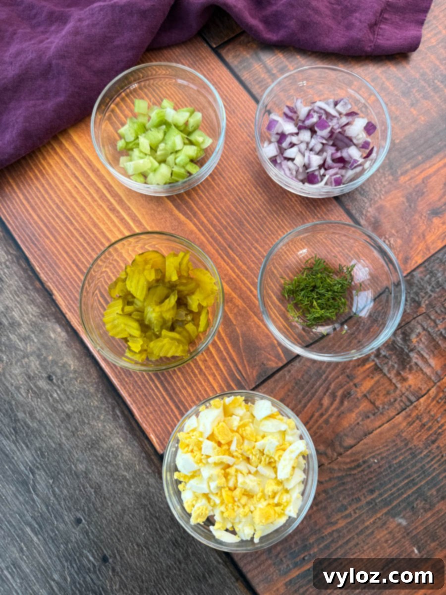 Dill-icious Creamy Potato Salad 3 A top-down view of five small glass bowls containing prepped ingredients for dill pickle potato salad. The bowls hold diced celery, chopped red onion, chopped dill pickles, fresh dill, and chopped hard-boiled eggs. They are placed on a wooden surface with a dark purple cloth in the background.