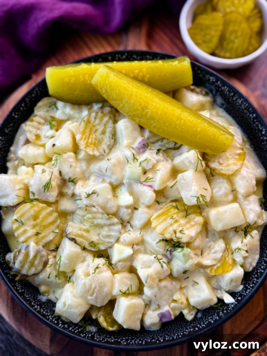 Dill-icious Creamy Potato Salad 9 A close-up image of a bowl of creamy dill pickle potato salad. The salad features cubed potatoes, diced pickles, red onion, celery, and fresh dill, all coated in a creamy dressing. The dish is garnished with sliced pickles and served in a black speckled bowl, with a background of a purple cloth and a small dish of pickles.