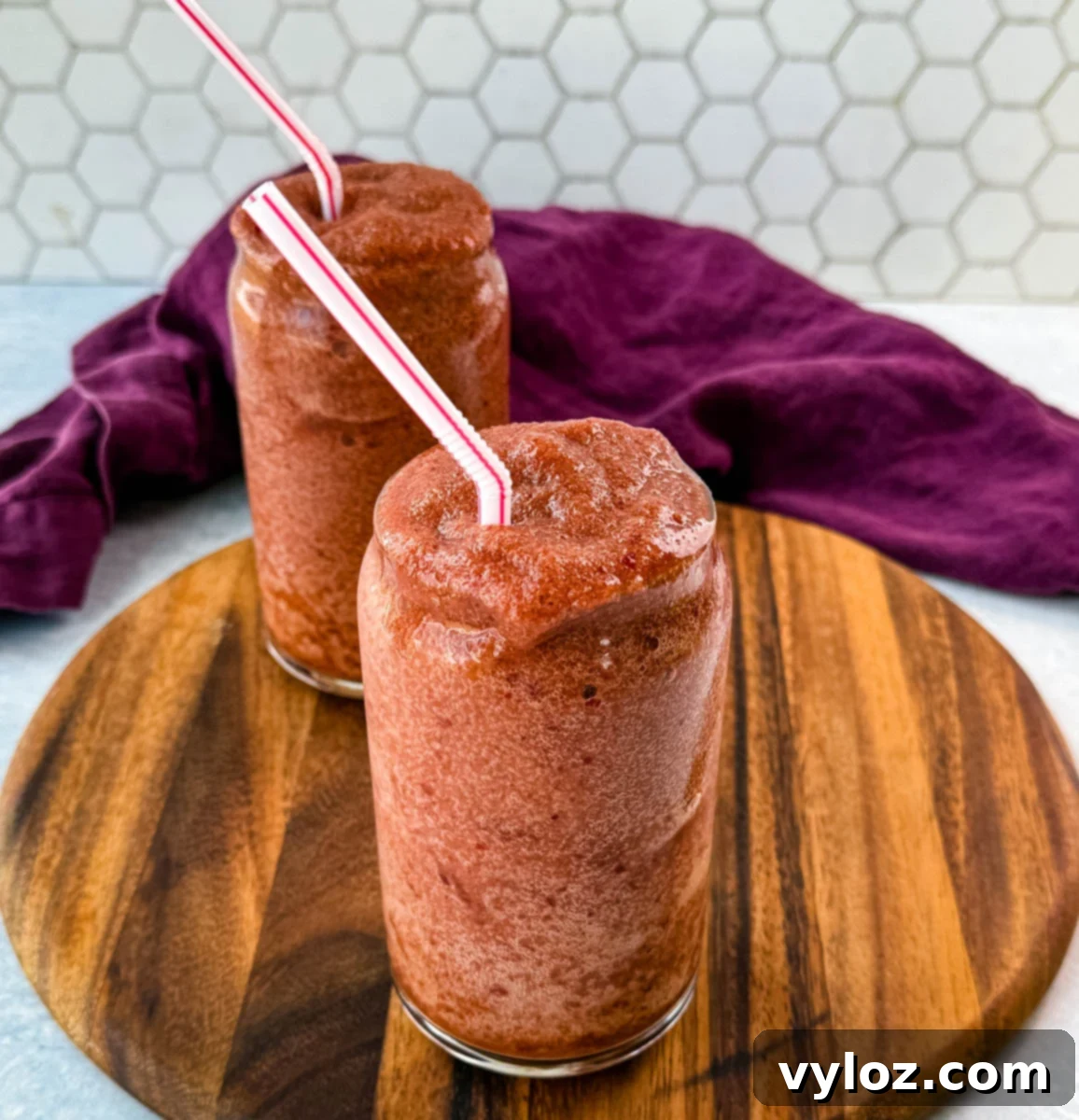 A close-up shot of two frosty cherry slushies with a rich red color and perfectly blended texture. The wooden board and purple cloth create a cozy, inviting atmosphere.